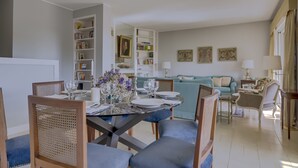 Un coin repas avec une table dressée pour quatre personnes, entourée de chaises. On aperçoit une bibliothèque sur la gauche et un canapé au fond de la pièce.