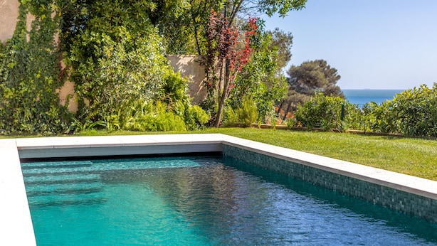 Une piscine dont le bord est revêtu de carrelage, entourée d’une végétation luxuriante et offrant une vue sur la mer.