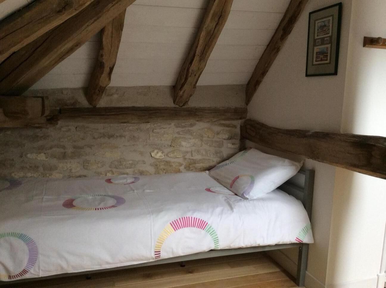 A single bed with a white mattress and colorful circular patterns, wooden beams, and a stone wall in a rustic room.