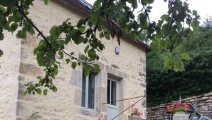 Una casa de piedra con una ventana pequeña, una puerta y una escalera que conduce a un muro de piedra. Hay plantas y flores alrededor de la casa.