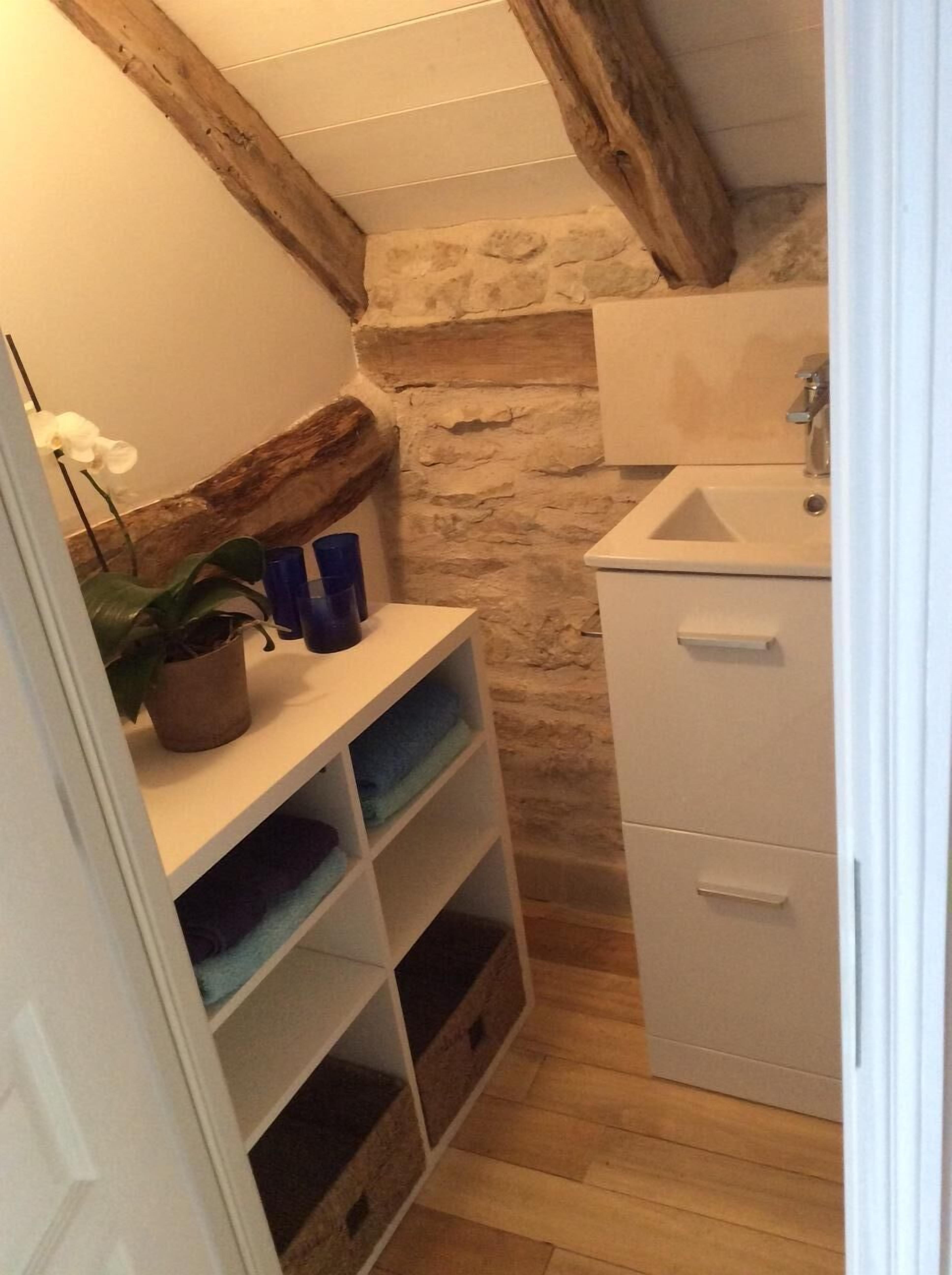 Une petite salle de bain avec un mur en pierre, des poutres en bois, un lavabo et des étagères où sont pliées des serviettes.