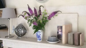 Una habitación con un reloj en la pared, un jarrón con flores, una lámpara, un adorno metálico y algunas velas sobre una estantería.