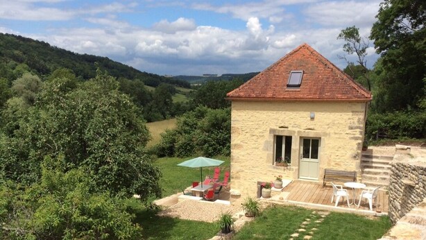 Una casa de piedra con tejado de baldosas, un patio con mobiliario exterior y vistas a un valle verde.