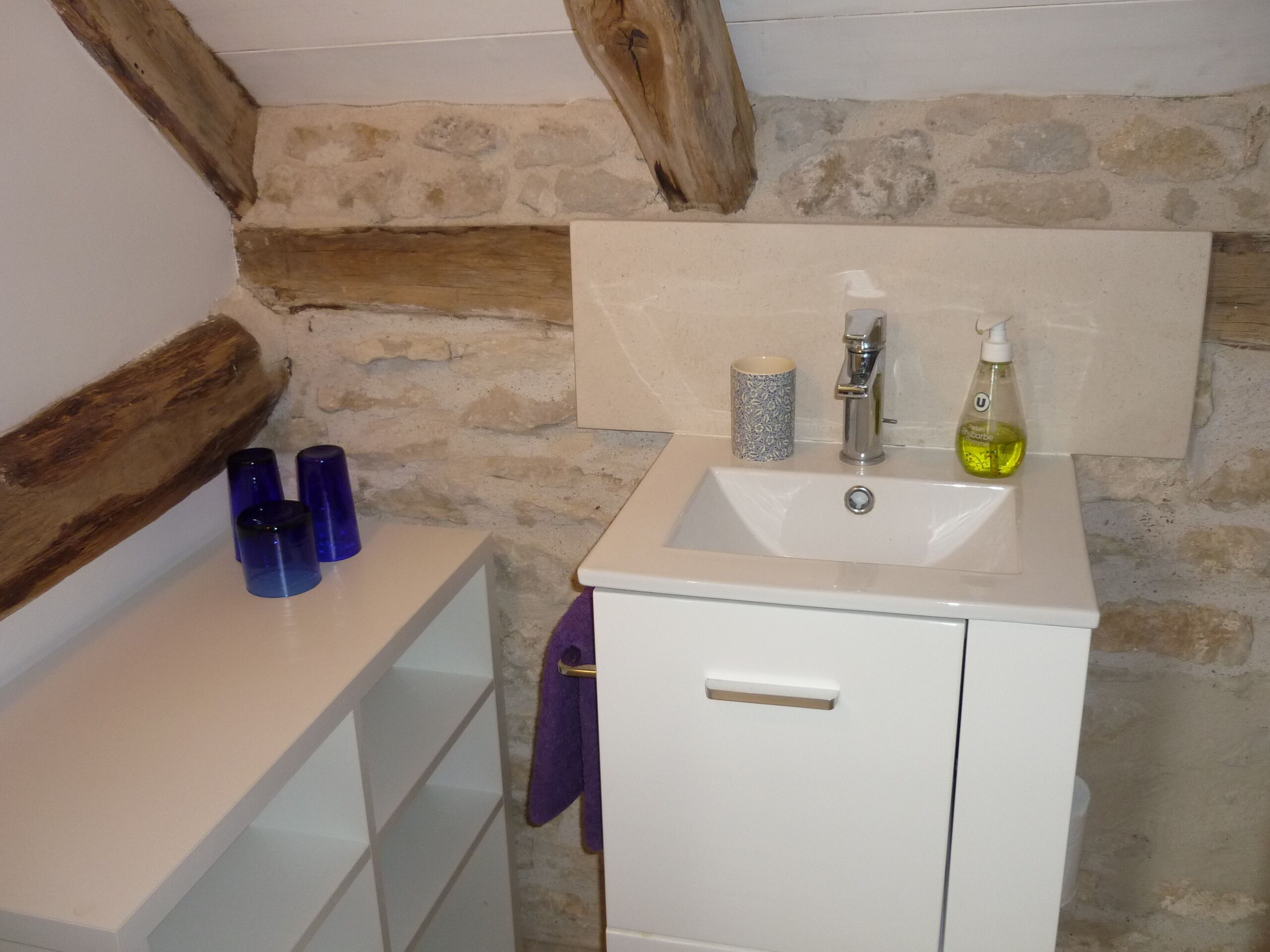 Une salle de bain avec un lavabo blanc, un miroir et un mur en pierre. On peut voir trois bouteilles bleues sur le comptoir.