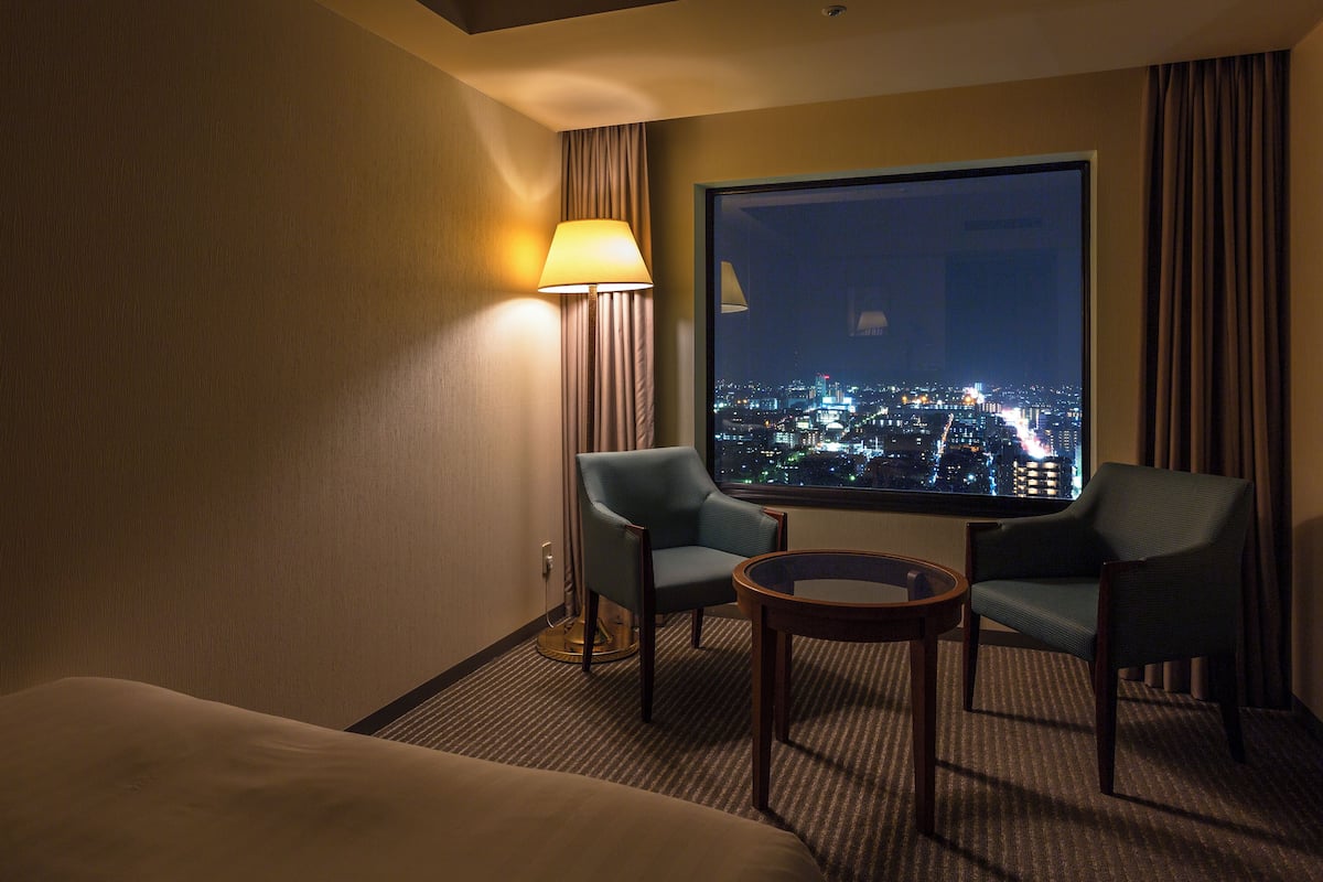 Ein Hotelzimmer mit großem Fenster, zwei Stühlen, einem kleinen Tisch und einer Lampe.