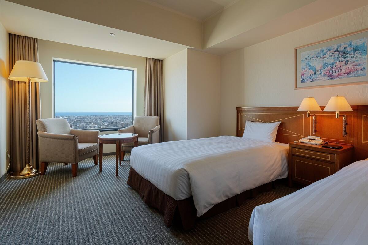 Ein Hotelzimmer mit zwei Betten, einer Sitzecke mit einem Sessel und einem kleinen Tisch, einem großen Fenster mit Blick und einem Gemälde an der Wand.