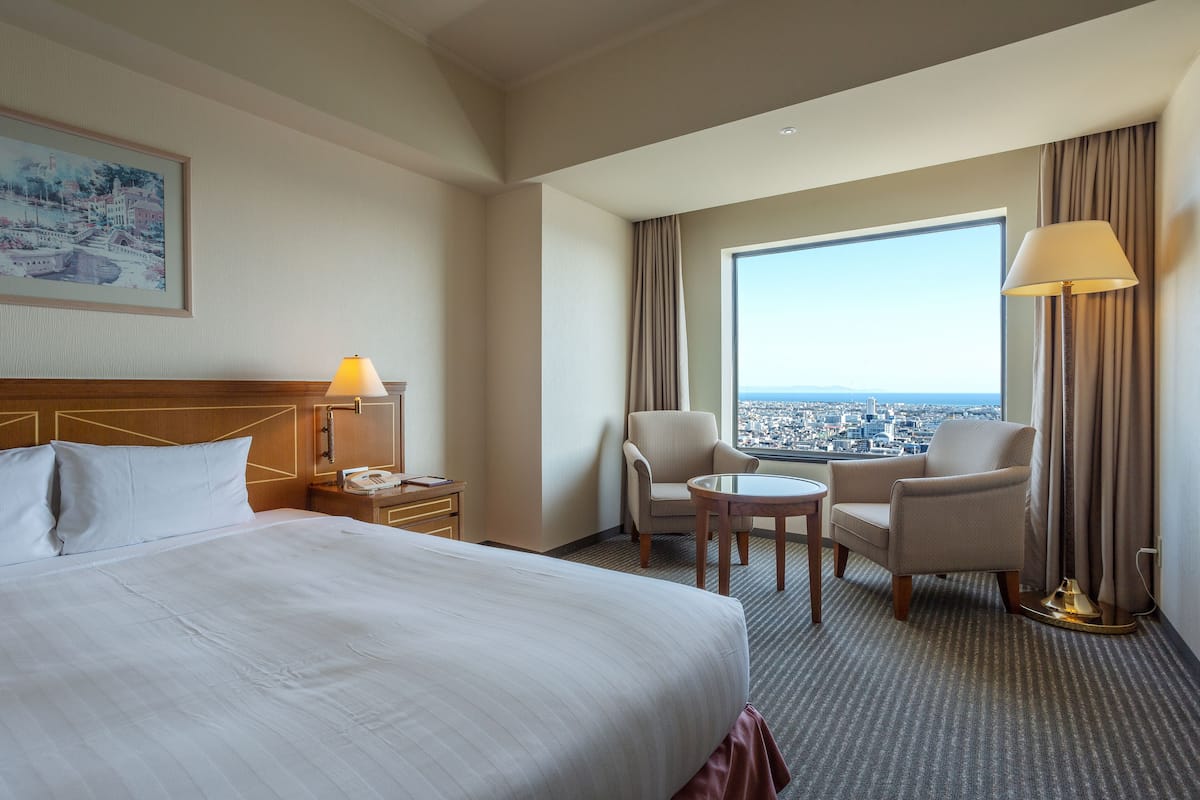 Ein Hotelzimmer mit einem großen Bett, einem Schreibtisch, zwei Stühlen, einer Lampe und einem großen Fenster mit Blick auf die Stadt.