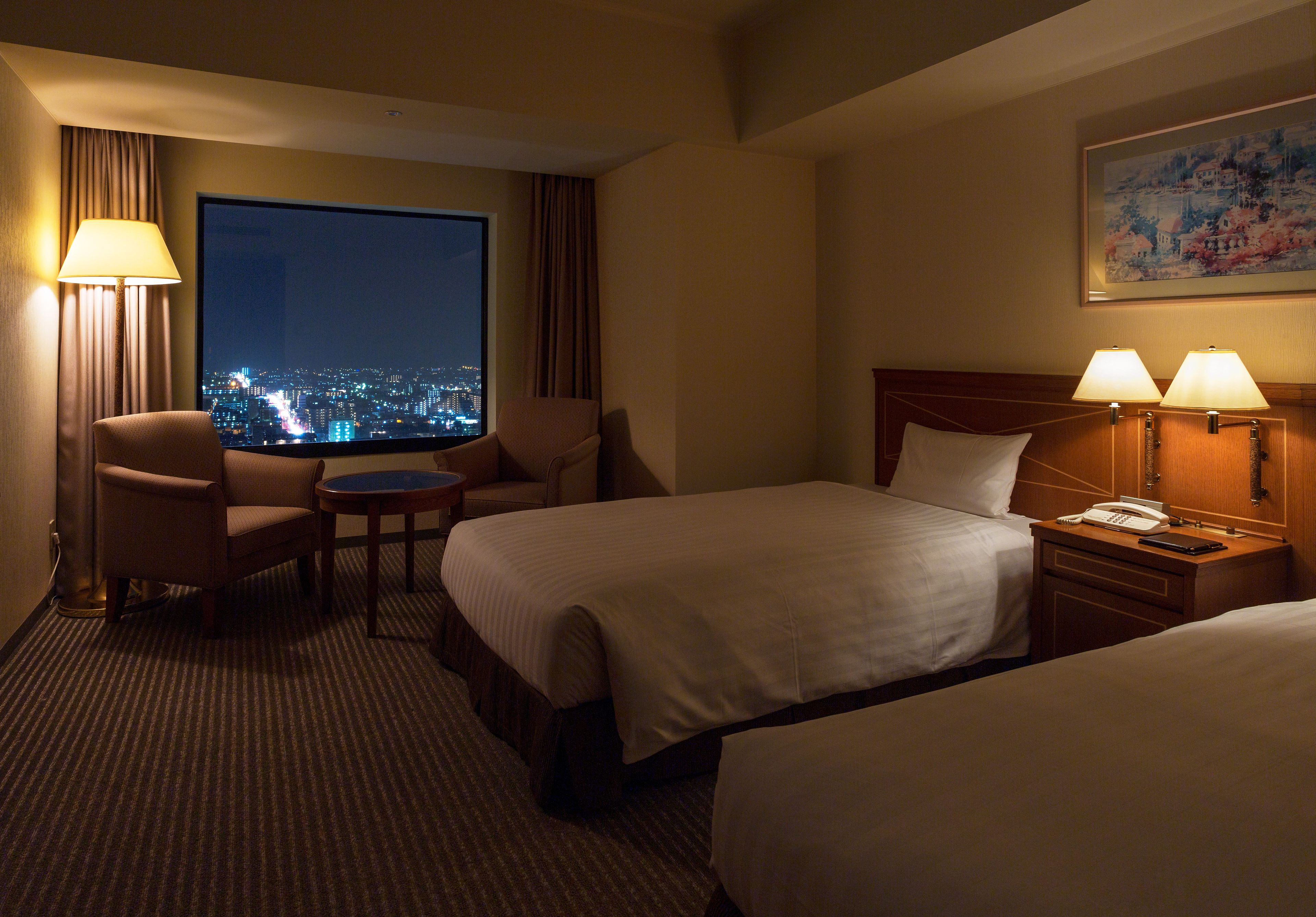 A hotel room with two beds, a desk, a chair, a lamp, and a large window with a city view.