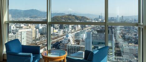 A room with two blue patterned chairs, a small round table, and a large window offering a cityscape view with mountains in the distance.