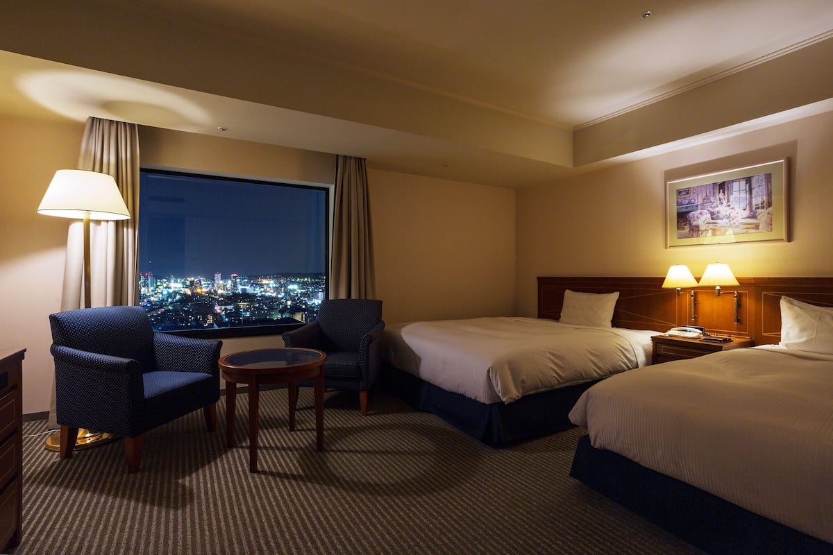 Ein Hotelzimmer mit zwei Betten, einem Schreibtisch, einem Stuhl, einer Lampe und einem großen Fenster mit Blick auf die Stadt.