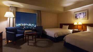 Ein Hotelzimmer mit zwei Betten, einem Schreibtisch, einem Stuhl, einer Lampe und einem großen Fenster mit Blick auf die Stadt.