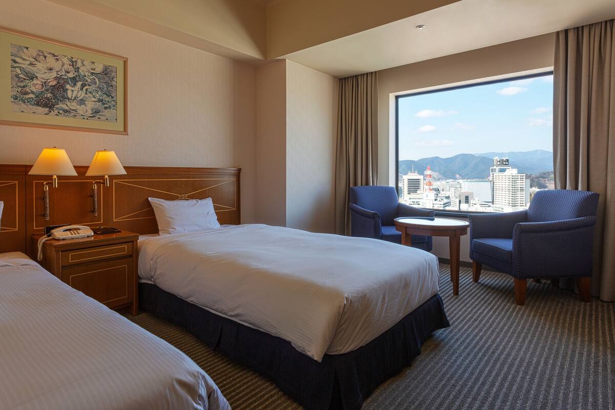 Ein Hotelzimmer mit zwei Betten, einem Schreibtisch mit Telefon und Blick auf eine Stadtlandschaft durch große Fenster.