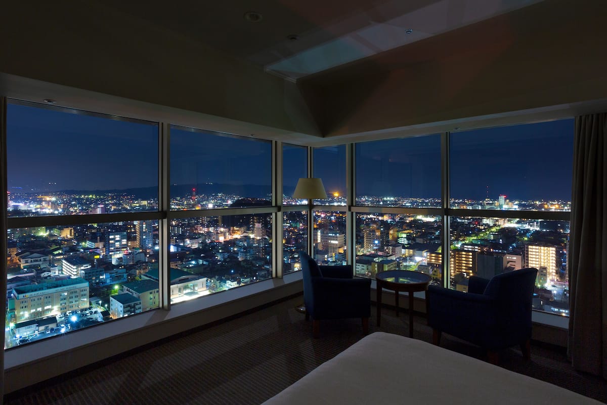 Ein Hotelzimmer mit großen Fenstern und Blick auf die beleuchtete Stadt bei Nacht.