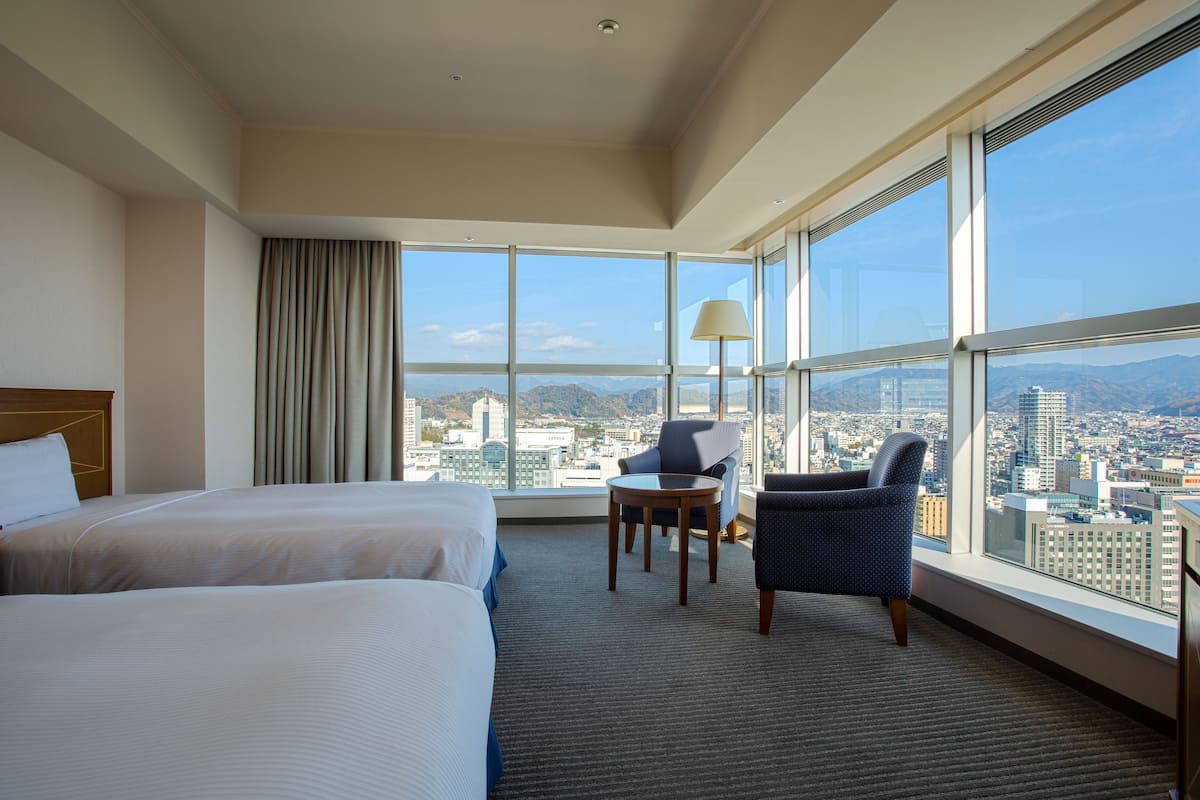 Ein Hotelzimmer mit zwei Betten, einem kleinen Tisch, zwei Stühlen, einer Lampe und großen Fenstern mit Blick auf die Stadt.