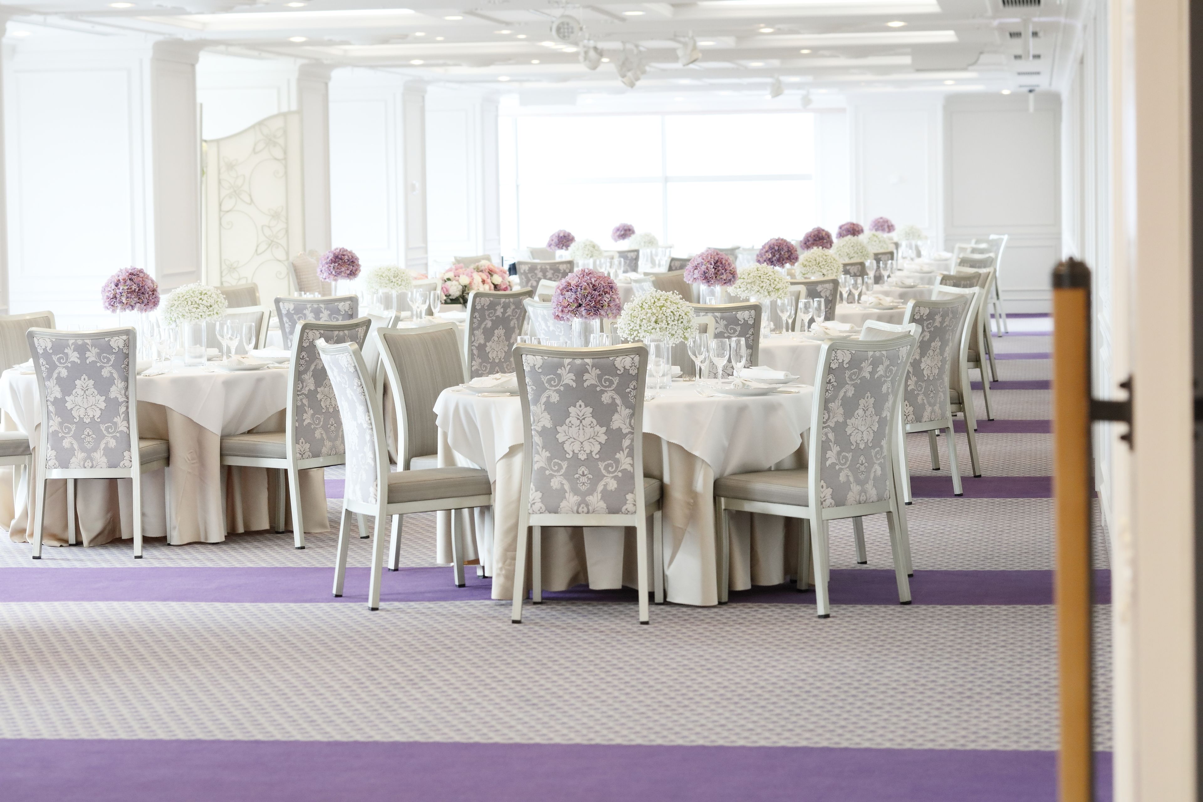 A banquet hall with rows of tables and chairs, each set with white tablecloths and floral centerpieces.