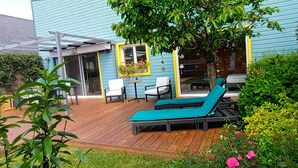 Una terraza de madera con una silla de descanso y una mesa, rodeada de vegetación y un edificio con fachada azul.