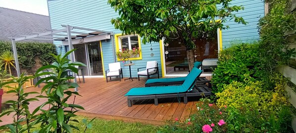 A wooden deck with a lounge chair and a table, surrounded by greenery and a building with a blue exterior. - Quiet new wood house Near beaches, Surf Spa Pool Terrace (Plomeur)