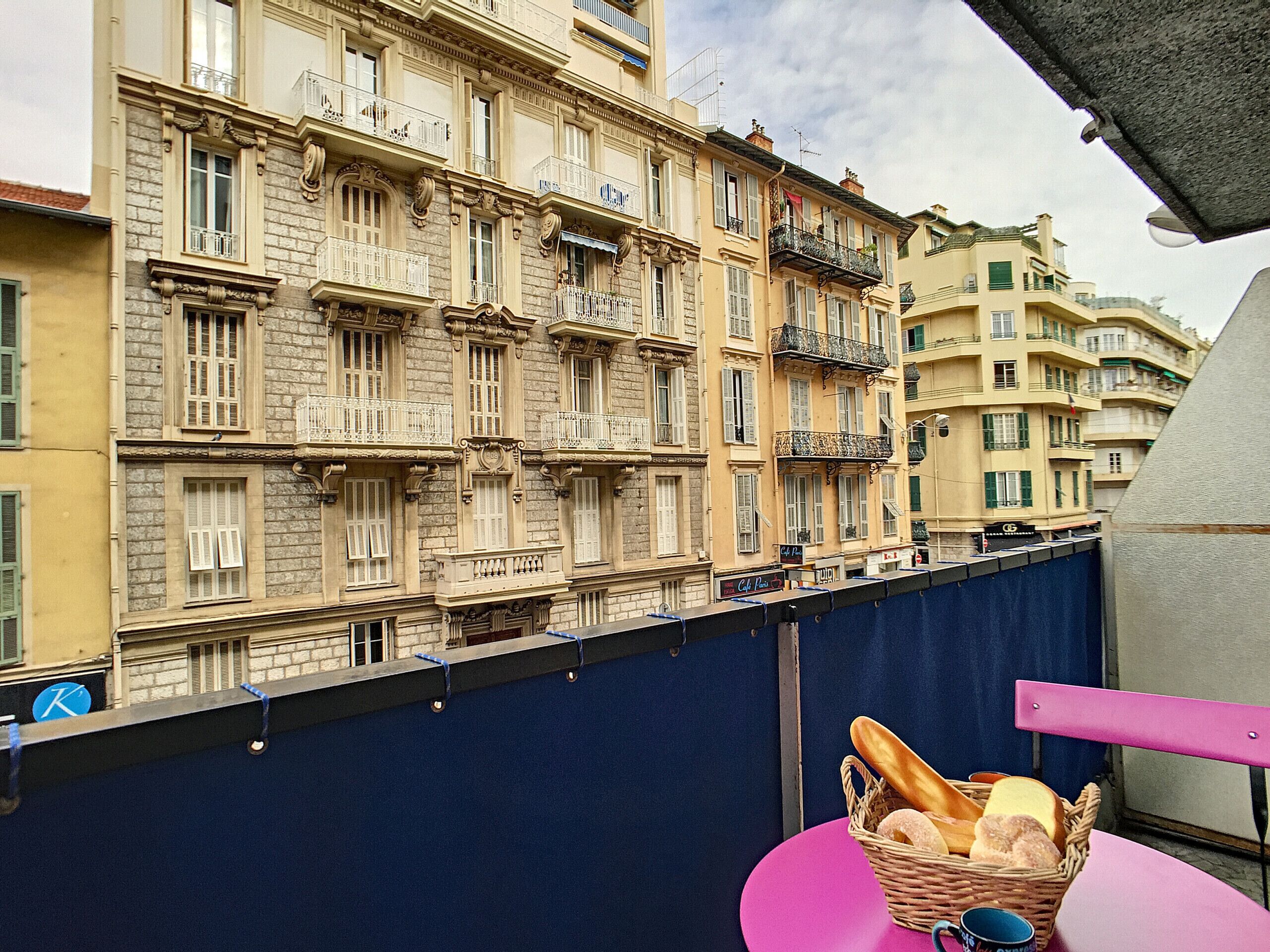 A balcony with a wicker basket containing bread and a mug, overlooking a row of historic buildings with ornate balconies and shutters.