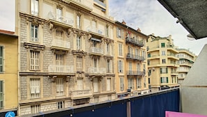 A balcony with a wicker basket containing bread and a mug, overlooking a row of historic buildings with ornate balconies and shutters.