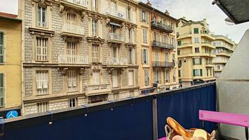 A balcony with a wicker basket containing bread and a mug, overlooking a row of historic buildings with ornate balconies and shutters.