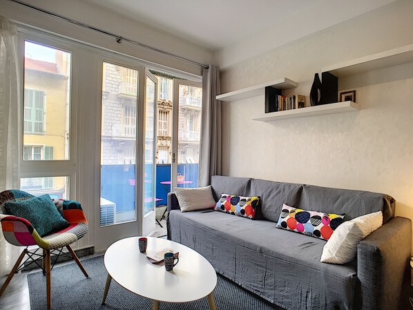 Een moderne woonkamer met een grijze bank, kleurrijke kussens, een klein wit tafeltje en een stoel met een gestreept kussen.
