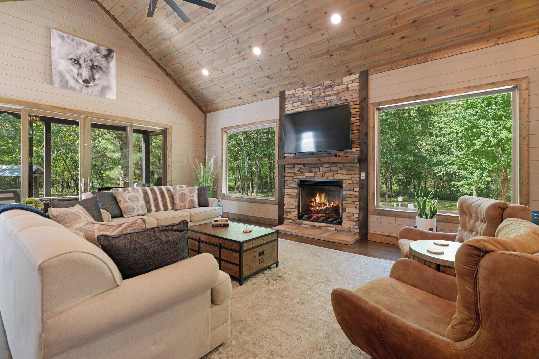 A cozy living room with a stone fireplace, comfortable seating, and large windows offering a view of the outdoors.