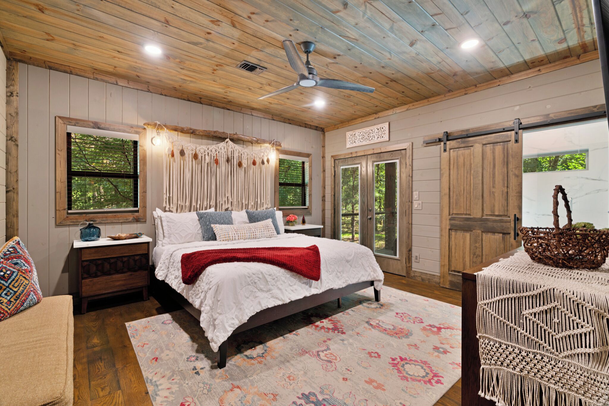A bedroom with a wooden ceiling, a bed with white bedding, a nightstand, a dresser, and a window with a view of trees.