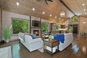 A spacious living room with a fireplace, large windows, and a kitchen area visible through an open door.