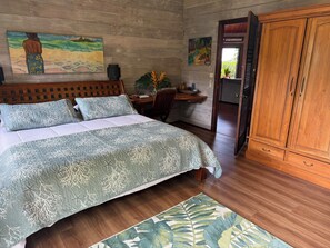 A bedroom with a bed, a wooden headboard, a nightstand, a desk, a cabinet, and two paintings on the wall. - Beautiful Private Home with Stunning Views of Opunohu Bay (Opunohu Bay)