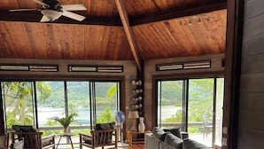 Une pièce spacieuse avec un plafond en bois, de grandes fenêtres donnant sur un paysage tropical, un canapé gris, des meubles en bois et un ventilateur.