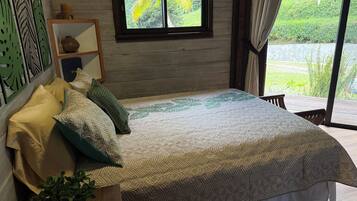 Un dormitorio con una cama grande, una ventana con vistas a zonas verdes, una mesita de noche con una planta y un adorno en la pared.