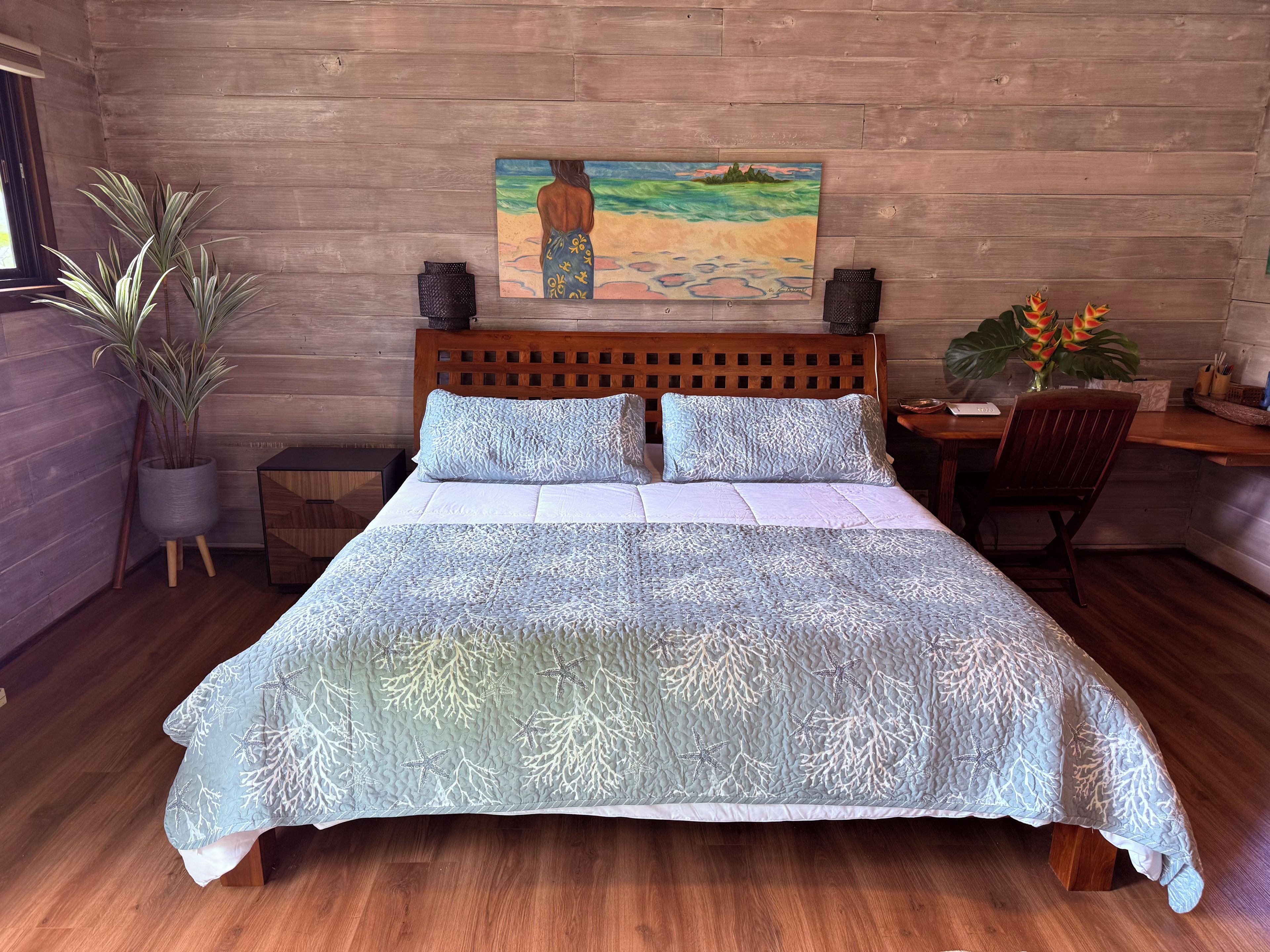 Un lit avec une couvre-lit bleue à motifs, une tête de lit en bois et un tableau représentant une scène de plage accroché au mur.