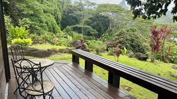A wooden deck with a bench and a table, overlooking a lush garden with various plants and a distant mountain.