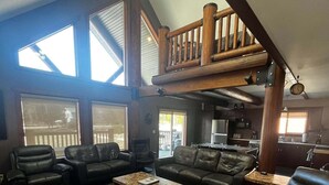 A spacious living room with a high ceiling, large windows, and a loft area. - Raven Cabin Vacation Rental (Valemount)
