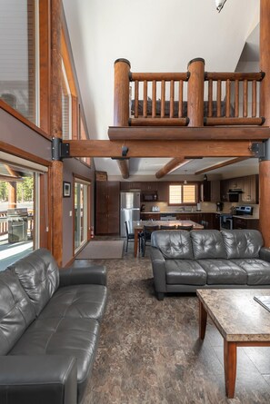 A spacious living area with a high ceiling, a balcony with wooden railings, and a kitchen in the background.