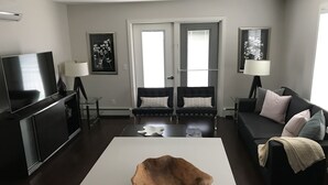 A modern living room with a flat-screen TV, a large white coffee table, and a black sofa with multiple cushions. There are two black chairs and a floor lamp. The room has a large window with blinds and a door leading to another room. - Comfortable and Spacious 2BD, 2BA New Built Condo (Saskatoon)