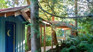A wooden cabin with a blue door featuring a crescent moon design, surrounded by dense forest and a wooden deck. - Westover Bed and Breakfasts (Salt Spring Island)