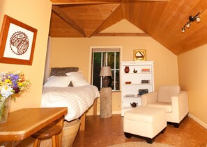 A cozy bedroom with a bed, a white armchair, a small table, and a bookshelf. - Westover Bed and Breakfasts (Salt Spring Island)