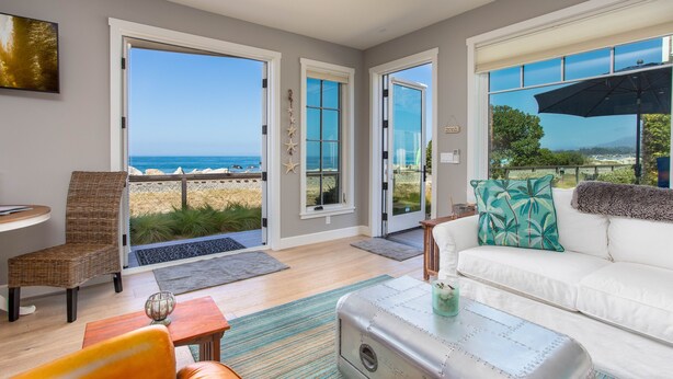 A spacious living area with a white sofa, a wicker chair, and a small table. Large glass doors open to a balcony with a view of the ocean. - Beach front Sea Loft in Santa Barbara.  Weekly discounts.  Best beach sunsets! (Carpinteria)