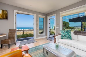A spacious living area with a white sofa, a wicker chair, and a small table. Large glass doors open to a balcony with a view of the ocean.