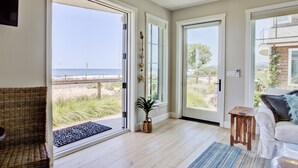A spacious room with large sliding glass doors leading to a beach view, a wicker chair, a small wooden table, and a white sofa. - Beach front Sea Loft in Santa Barbara.  Weekly discounts.  Best beach sunsets! (Carpinteria)