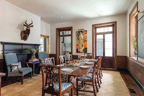 A dining area with a long wooden table set for six, surrounded by upholstered chairs. A mounted deer head is on the wall, and a colorful painting hangs above the table. - Casa Zoë | Grand Sparrow House (Charleston)