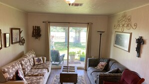 A living room with two sofas, a coffee table, and a red armchair. There is a large window with curtains, a ceiling fan, and wall-mounted artwork.