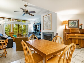 A spacious living room with a wooden dining table, comfortable seating, a fireplace, and a large window overlooking greenery.