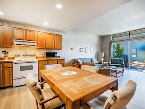 A spacious kitchen with wooden cabinets, a dining table, and chairs. There is a microwave, stove, and a large window offering a view of the outdoors.