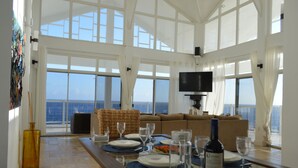 A dining area with a table set for four, large windows offering a view of the sea, a television mounted on the wall, and a large painting on the wall. - The Most Beautiful and Relaxing Oceanfront Views of Curacao (Santa Martha)