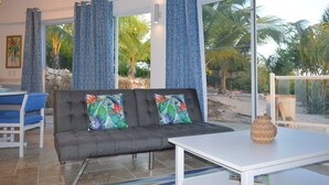 A modern living room with a grey sofa, colorful pillows, a white table, and large windows with blue patterned curtains. - The Most Beautiful and Relaxing Oceanfront Views of Curacao (Santa Martha)