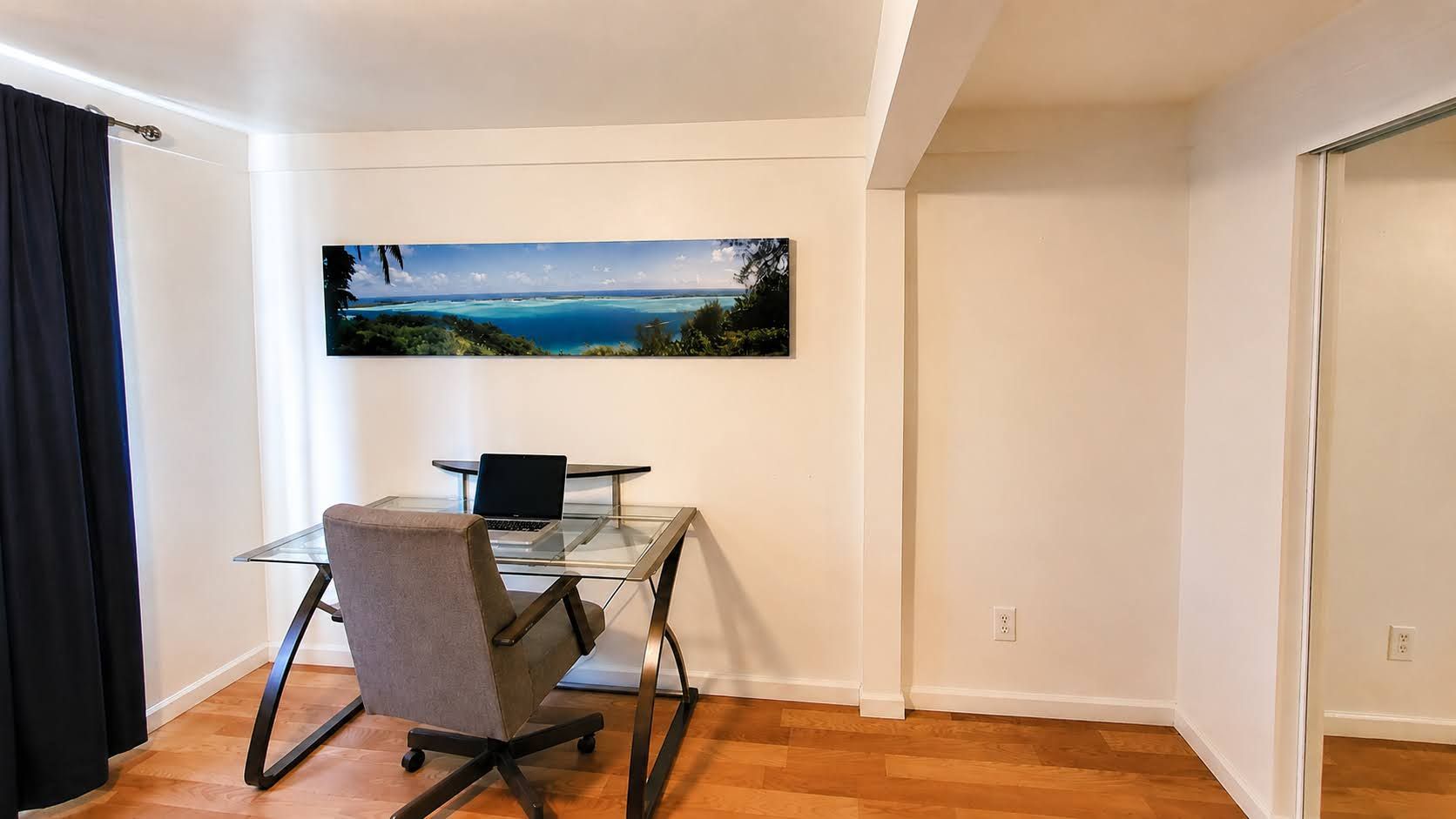 A room with a glass desk, a laptop, a chair, and a painting of a landscape.