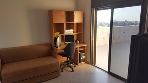 Ein Zimmer mit einem braunen Sofa, einem Holztisch mit einem Computer und einer Glastür zum Balkon.