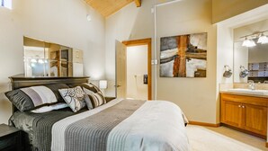 Een slaapkamer met een bed, nachtkastje, houten plafond, schilderij en een badkamer met wastafel en kastjes.